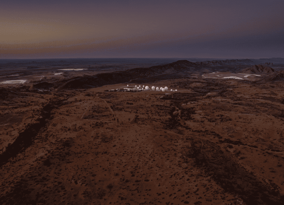 The radome spheres of Pine Gap intelligence facility visible against the Australian desert sky at dusk, small against the vast red landscape