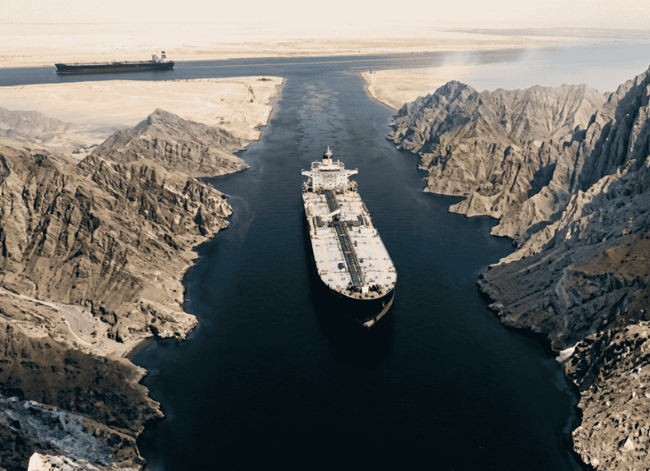 A large oil tanker transits the Strait of Hormuz through the narrow navigable channel, desert terrain visible on both sides, conveying the strategic vulnerability of the world’s most critical energy chokepoint