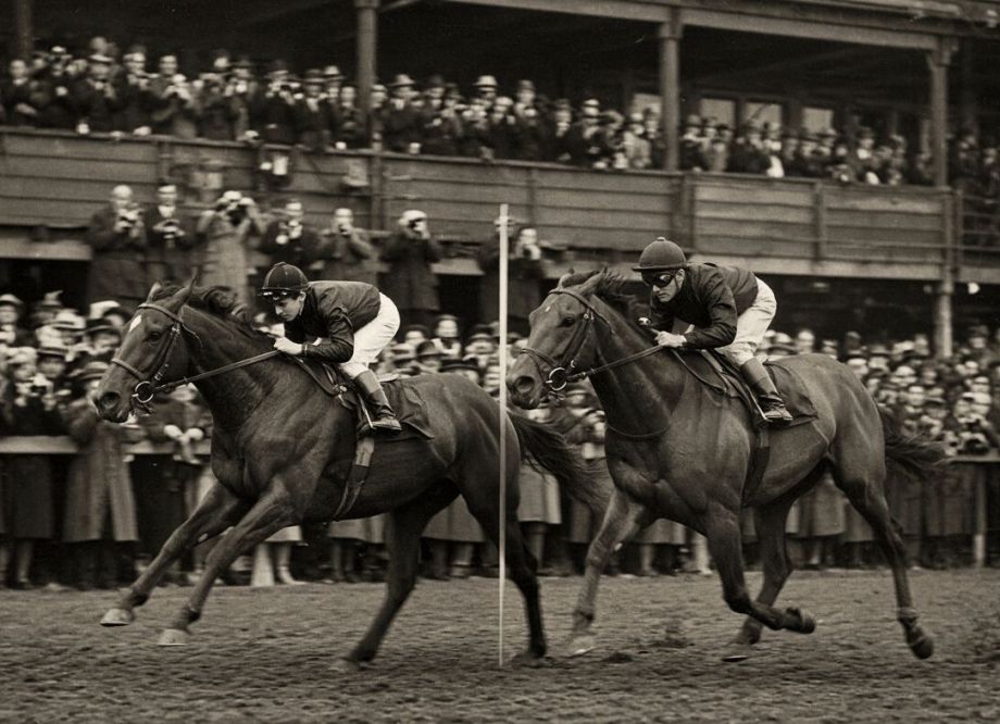 A 1940s-style black-and-white racing photograph. Two horses gallop right to left past a white finishing post, the leader about a length clear, watched by a packed wooden grandstand of men in hats, many holding cameras and binoculars.